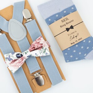 May include: A light blue suspender set with a floral bow tie and a pair of blue polka dot socks. The socks have a tag that reads "It's time to Suit up!!" and the date August 12, 2023. The suspenders have silver clips and a gray leather patch.