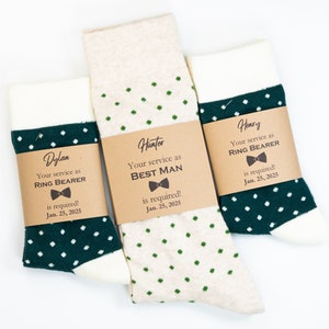 May include: Three pairs of socks with brown tags. The socks are white with green polka dots. The tags have the names Dylan, Hunter, and Henry and the text "Your service as Ring Bearer is required! Jan. 25, 2025" or "Your service as Best Man is required! Jan. 25, 2025".