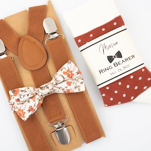 Puede incluir: Un conjunto de tirantes marrones con una pajarita floral y un par de calcetines blancos con lunares rojos. Los calcetines tienen una etiqueta que dice "Mason Ring Bearer Oct. 21, 2023".