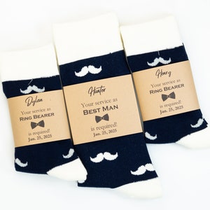 May include: Three pairs of navy blue socks with white mustache designs and brown paper tags. The tags have the names "Dylan", "Hunter", and "Henry" and the text "Your service as RING BEARER is required! Jan. 25, 2025" or "Your service as BEST MAN is required! Jan. 25, 2025".