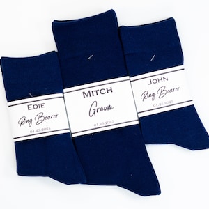 May include: Three pairs of dark blue socks with white labels. The labels have the names "Edie", "Mitch", and "John" and the title "Ring Bearer" or "Groom". The date "01.25.2025" is printed on each label.