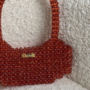 May include: A small, orange beaded handbag with a top handle. The bag has a gold label that reads "Knitkos".