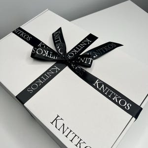 May include: A white gift box with a black ribbon tied in a bow. The ribbon has the word "KNITKOS" printed on it in a silver font.