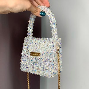May include: A small, white beaded handbag with a gold chain strap and a gold nameplate that reads "K.niikos".