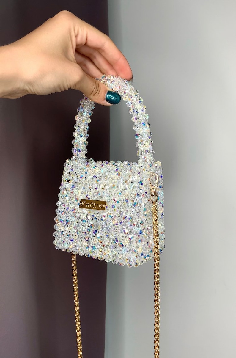 May include: A small, white beaded handbag with a gold chain strap and a gold nameplate that reads "K.niikos".
