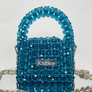 May include: A small, teal blue beaded handbag with a top handle and a silver chain strap. The bag has a rectangular shape and is made of small, faceted beads. The bag has a silver label with the word "Knitkos" on it.
