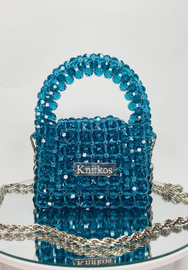 May include: A small, teal blue beaded handbag with a top handle and a silver chain strap. The bag has a rectangular shape and is made of small, faceted beads. The bag has a silver label with the word "Knitkos" on it.