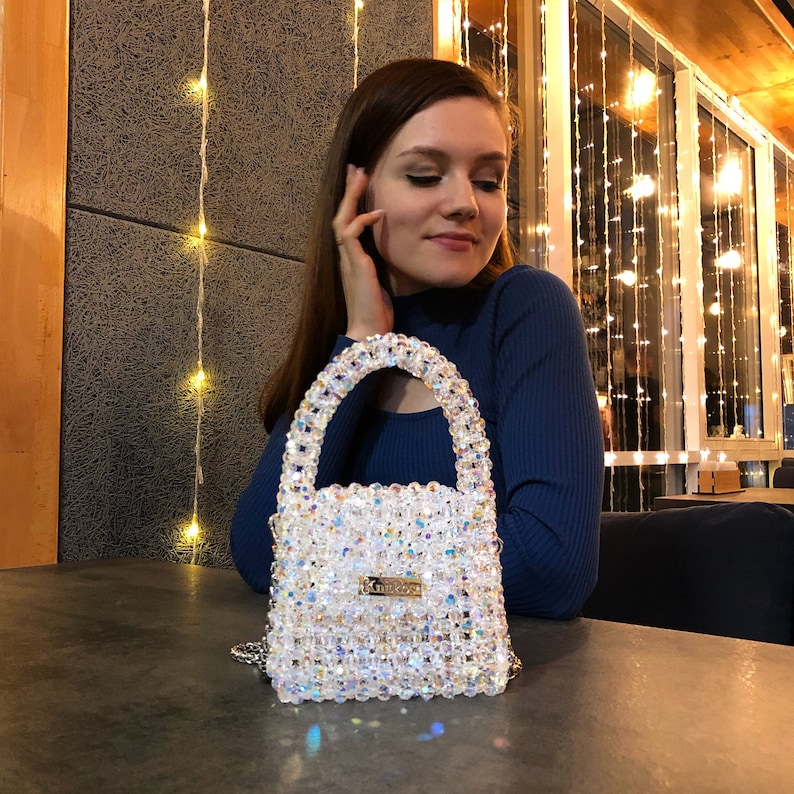 May include: A small, clear, iridescent beaded handbag with a gold clasp and a silver chain strap. The bag is made of many small, round beads that are strung together to create a unique and eye-catching design.