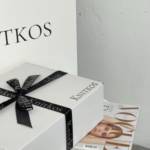 May include: A white gift box with a black ribbon and the word "Knitkos" printed on the top and sides. The box is sitting on a white table with a copy of Vogue magazine on top.