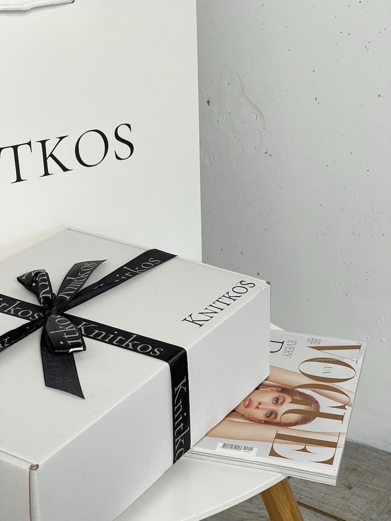 May include: A white gift box with a black ribbon and the word "Knitkos" printed on the top and sides. The box is sitting on a white table with a copy of Vogue magazine on top.