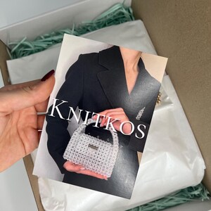 May include: A white beaded handbag with a gold handle. The bag is being held by a person wearing a black blazer. The text "KNITKOS" is visible on the card.