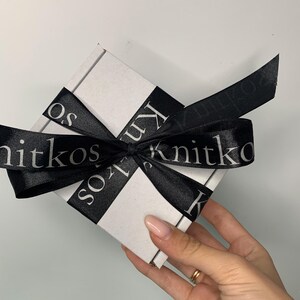 May include: A white gift box with a black ribbon tied around it. The ribbon has the text "Knitkos" printed on it in white.