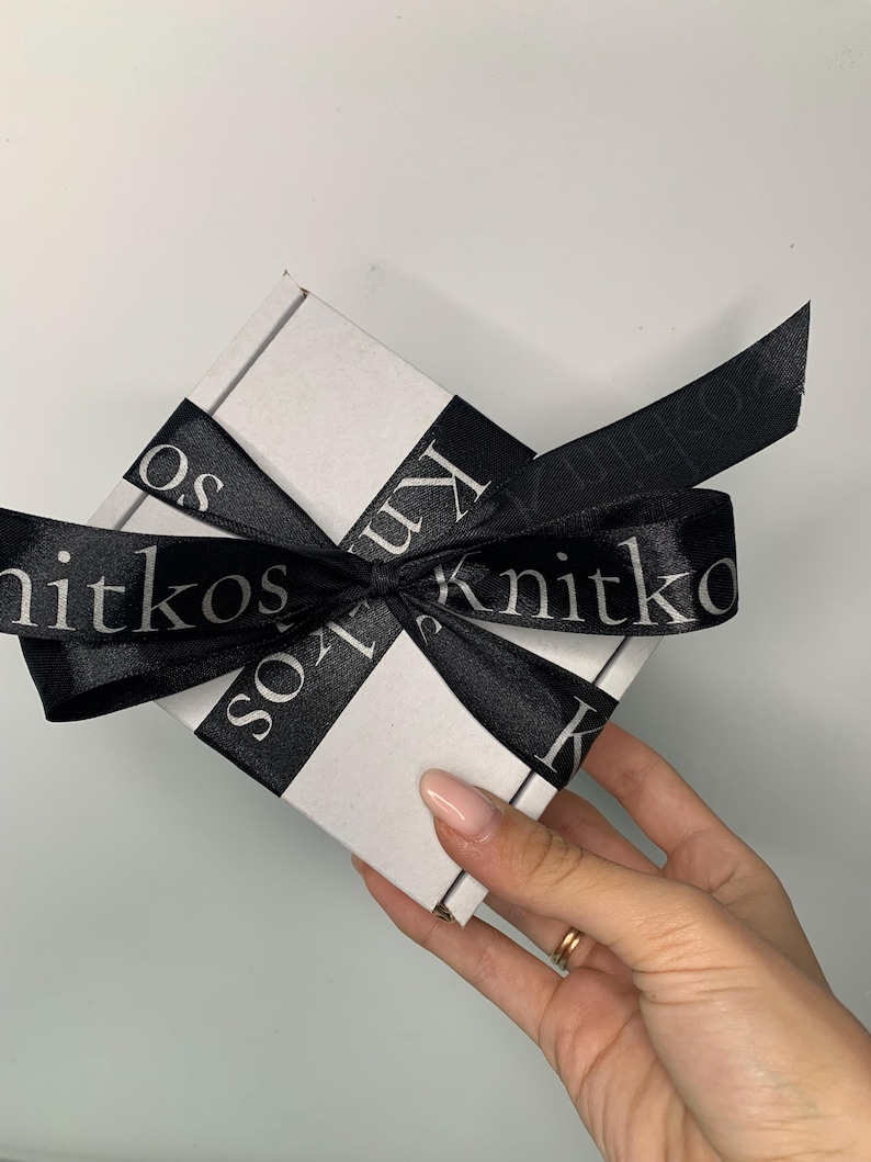 May include: A white gift box with a black ribbon tied around it. The ribbon has the text "Knitkos" printed on it in white.
