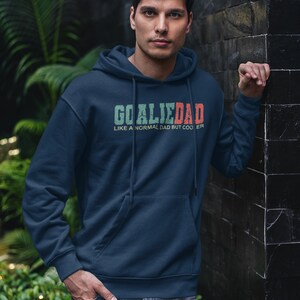 K&ouml;nnte beinhalten: Ein marineblauer Hoodie mit einem wei&szlig;en Kordelzug. Der Hoodie hat eine gr&uuml;ne und rote Grafik mit der Aufschrift "Goalie Dad Like a normal dad but cooler."