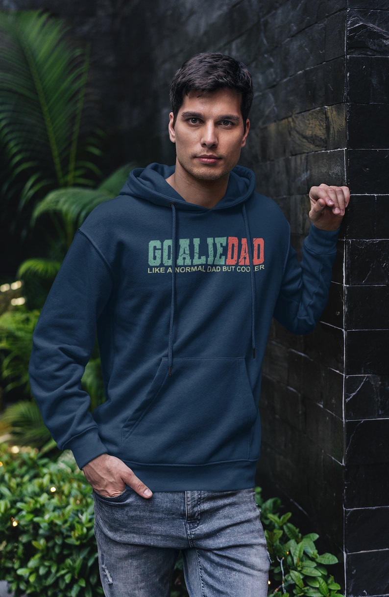 K&ouml;nnte beinhalten: Ein marineblauer Hoodie mit einem wei&szlig;en Kordelzug. Der Hoodie hat eine gr&uuml;ne und rote Grafik mit der Aufschrift "Goalie Dad Like a normal dad but cooler."