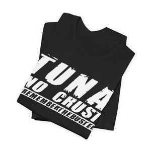 Pu&ograve; includere: Una t-shirt nera con testo bianco in stile consumato che recita TUNA NO CRUST REMEMBER THE BUSTER. La maglietta &egrave; piegata ordinatamente, mostrando la stampa grafica in modo prominente sulla parte anteriore contro il tessuto nero uniforme.