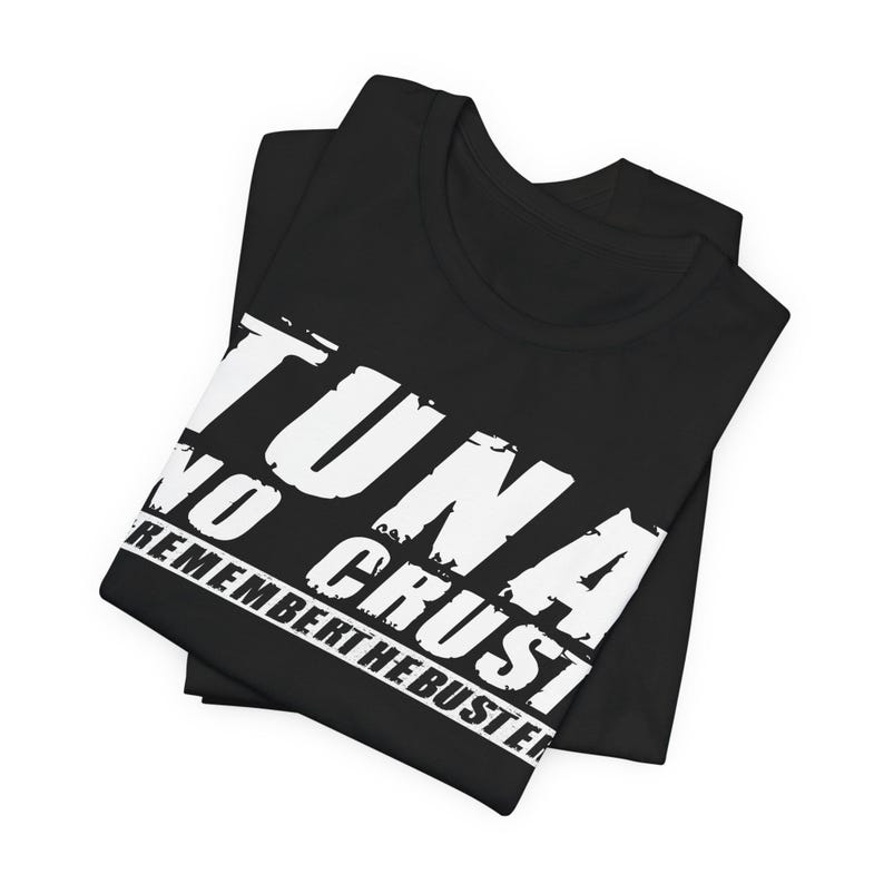 Pu&ograve; includere: Una t-shirt nera con testo bianco in stile consumato che recita TUNA NO CRUST REMEMBER THE BUSTER. La maglietta &egrave; piegata ordinatamente, mostrando la stampa grafica in modo prominente sulla parte anteriore contro il tessuto nero uniforme.