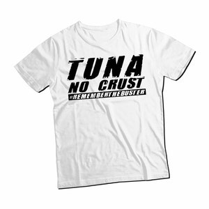 Pu&ograve; includere: Una t-shirt bianca a maniche corte con testo nero stampato sul petto che recita TUNA NO CRUST e #REMEMBERTHEBUSTER. La grafica utilizza un carattere in grassetto dall'aspetto usurato su un capo dotato di un classico scollo rotondo.