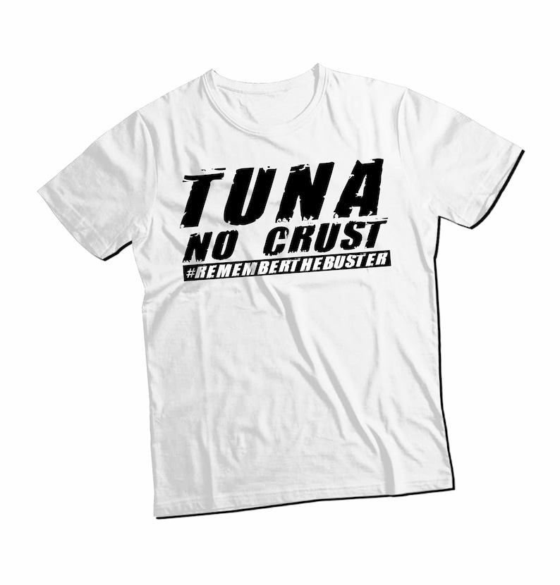 Pu&ograve; includere: Una t-shirt bianca a maniche corte con testo nero stampato sul petto che recita TUNA NO CRUST e #REMEMBERTHEBUSTER. La grafica utilizza un carattere in grassetto dall'aspetto usurato su un capo dotato di un classico scollo rotondo.