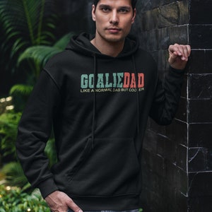 K&ouml;nnte beinhalten: Ein schwarzer Hoodie mit einem wei&szlig;en, gr&uuml;nen und roten Grafikdruck, auf dem "Goalie Dad Like a normal dad but cooler" steht.