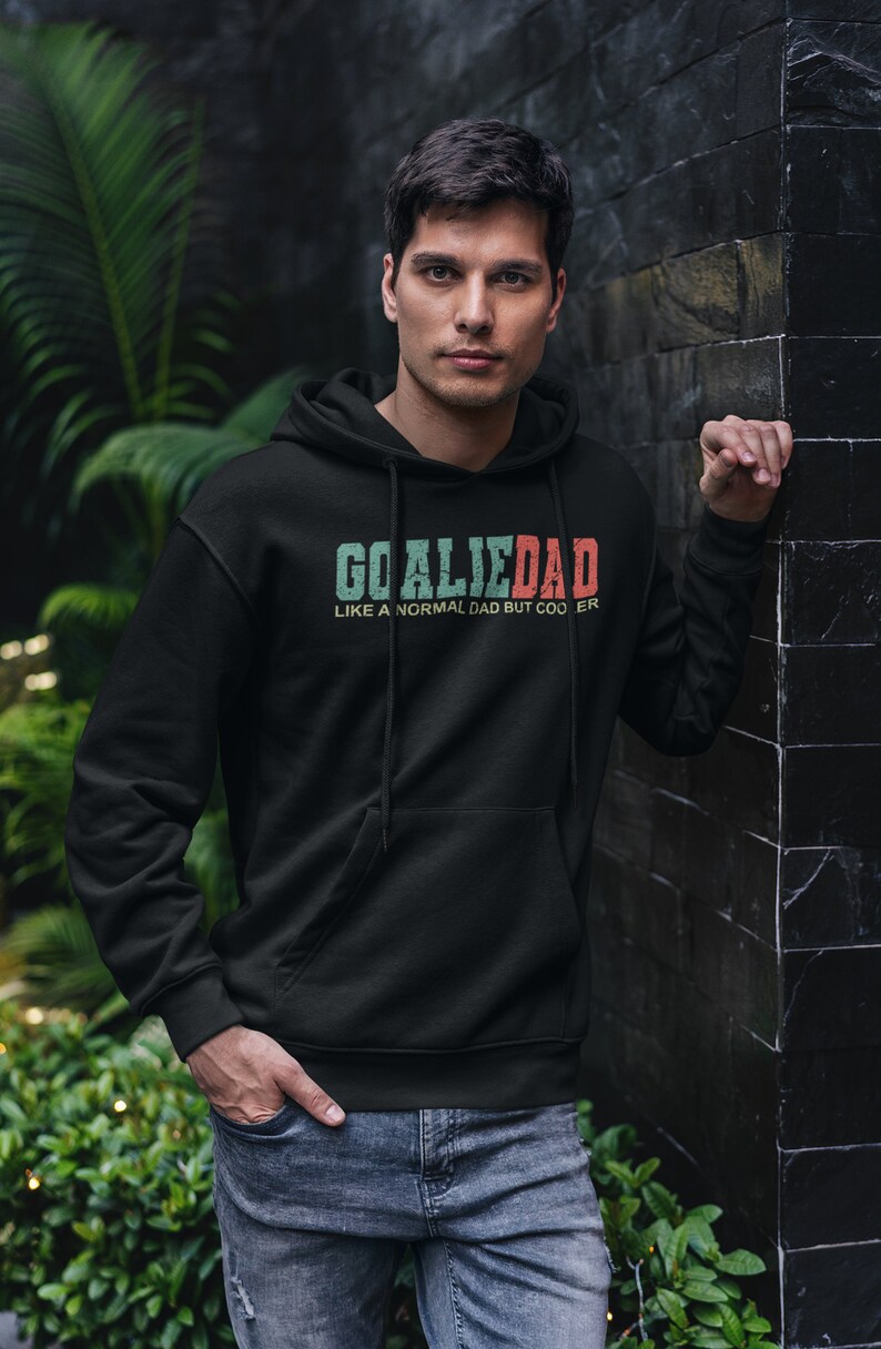 K&ouml;nnte beinhalten: Ein schwarzer Hoodie mit einem wei&szlig;en, gr&uuml;nen und roten Grafikdruck, auf dem "Goalie Dad Like a normal dad but cooler" steht.