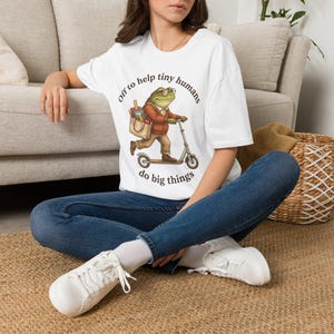 May include: A white short-sleeved t-shirt featuring a graphic of a frog wearing glasses and a brown jacket, riding a scooter while carrying a tote bag. The text "Off to help tiny humans do big things" is printed in a circular arc around the frog illustration.
