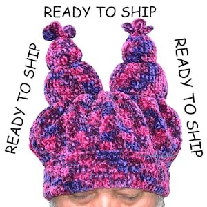May include: A handmade, crocheted hat in shades of pink and purple. The hat features a crown-like design with two tall, rounded peaks and a textured, plush appearance. The words "READY TO SHIP" are printed around the hat.