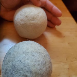 May include: Two wool dryer balls, one light beige and one gray, sit on a wooden surface. A hand gently touches the beige ball. These reusable dryer balls help reduce drying time and soften clothes.