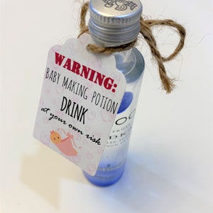 Puede incluir: Una pequeña botella de licor transparente con una tapa plateada y un lazo de cuerda marrón. Una etiqueta blanca con lunares rosas y una ilustración de un bebé rosa cuelga de la botella. La etiqueta dice "ADVERTENCIA: POTIÓN PARA HACER BEBÉS BEBE a tu propio riesgo".