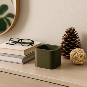 Peut inclure: Un récipient carré vert olive est posé sur une surface en bois clair, à côté d'une pile de livres et d'une paire de lunettes noires. Une pomme de pin et une boule tissée sont également sur la surface. Le récipient est vide.