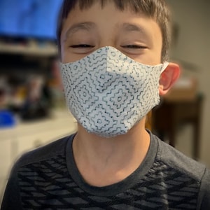 Puede incluir: Una persona joven usando una mascarilla facial blanca con un patrón geométrico azul. La mascarilla cubre la boca y la nariz. La persona sonríe y lleva una camisa gris oscura con un patrón blanco y negro.