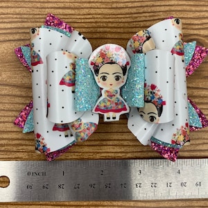 May include: A white bow with pink and blue glitter accents features a printed image of Frida Kahlo. The bow is approximately 4 inches wide.