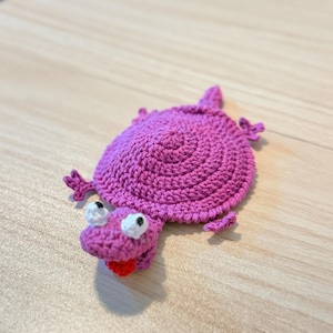 May include: A purple crocheted turtle-shaped coaster featuring a circular shell, four small legs, and a head with white eyes and a red tongue. The yarn texture is visible throughout the piece, showcasing a handmade construction with distinct stitches.