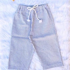 May include: Blue and white striped cropped pants with a drawstring waist. The pants are made of a lightweight fabric and have a relaxed fit. The drawstring is white and has decorative tassels. Three spools of thread are in the background.