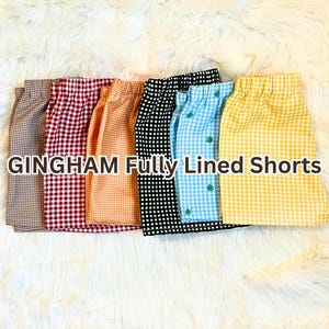 May include: A collection of fully lined gingham shorts in various colors, including brown, red, orange, black, blue, and yellow. The shorts are arranged in a row, showcasing their different patterns and hues. The text "GINGHAM Fully Lined Shorts" is displayed.