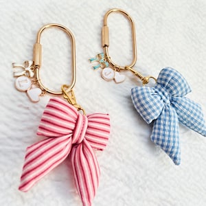 May include: Two keychains with gold-colored hardware. One has a pink and white striped bow, the other a blue and white gingham bow. Each keychain has charms, including hearts and letters. The keychains are on a white surface.