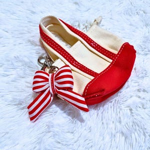 May include: A miniature canvas tote bag with red accents and a red and white striped bow keychain. The bag is off-white with red trim and a red base. The bow is attached with a silver clasp.