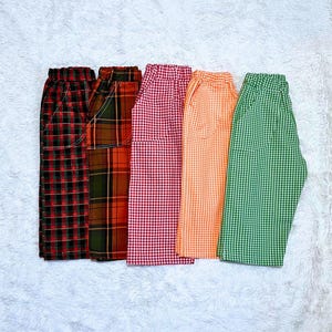 May include: Five pairs of casual pants in various plaid and gingham patterns. The colors include red, black, green, orange, and brown. The pants are folded and displayed on a white, textured surface.