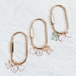 May include: Three gold-colored keychains with oval-shaped clasps. Each keychain has charms, including white hearts, bows, and circular pendants with letters. One keychain has a blue bow, and another has a pink bow.