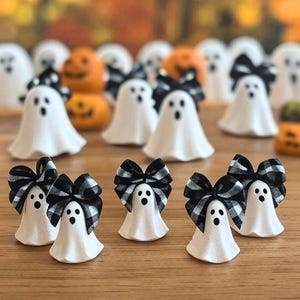 May include: White ghost figurines with black and white plaid bows. The ghosts have three black dot eyes and a black open mouth. Orange pumpkin figurines are in the background. The figurines are on a wooden surface.