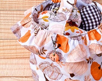 May include: A white fabric tote bag with a pumpkin pattern, featuring orange, tan, and gray pumpkins and leaves. The bag has a ruffled edge and a pumpkin charm attached. Inside are various fabric pumpkins.