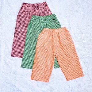 May include: Three pairs of checkered pants in red, green, and orange. The pants have a wide-leg design and pockets. The fabric has a classic gingham pattern, perfect for casual wear.