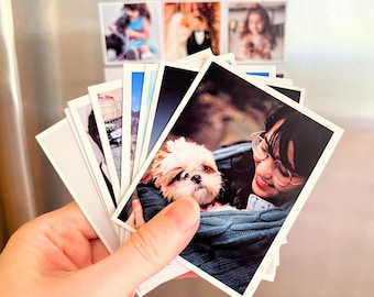 May include: A stack of square printed photos, held in a hand, showing a woman with glasses holding a small white dog. The photos are printed on white paper with a white border.