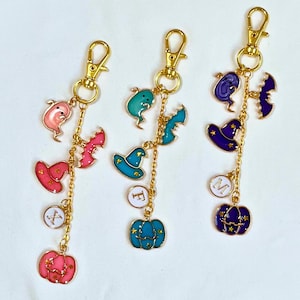 May include: Three Halloween-themed keychains with gold-colored clasps and chains. Each keychain features a series of charms, including ghosts, bats, witch hats, and pumpkins in pink, teal, purple, and blue. Some charms have letters.