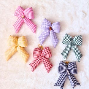 May include: Six gingham fabric bows in various colors: pink, yellow, red, green, purple, and navy blue. Each bow has a key ring attachment. The bows are arranged on a white, fluffy surface.