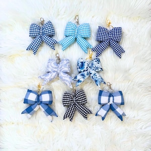 May include: Collection of fabric bow keychains in various blue and white patterns, including gingham and floral designs. Each bow is attached to a metal clasp. The keychains are arranged on a fluffy white surface.