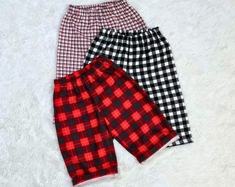 May include: Three pairs of plaid shorts in red and black, black and white, and red and pink. The shorts are arranged on a white, fluffy surface. The shorts have an elastic waistband.