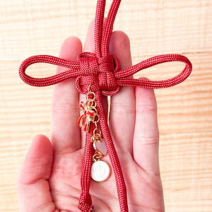May include: A red decorative knot with dangling charms. The knot is made of thick, woven material and features a bow, heart, and circular charm. The charms are gold-colored with red and white accents. The background is a light wood grain.