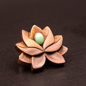 Pode incluir: Uma flor de lótus de madeira artesanal com pétalas detalhadas. A flor é de uma cor castanha quente e apresenta uma pequena pedra redonda verde clara no centro. Provavelmente um artigo decorativo ou um componente de joalharia.