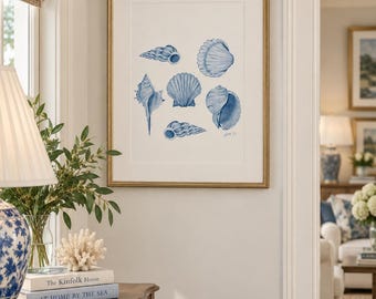 May include: A framed wall art print featuring a collection of six blue watercolor illustrations of various seashells arranged on a white background. The artwork is enclosed in a thin gold frame and includes a signature in the bottom right corner.